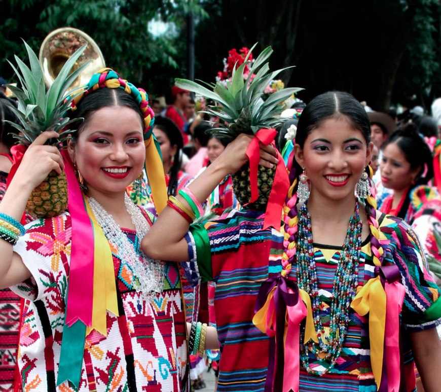 Two young women participants, each dressed in colorful dresses and carrying pineapples on their shoulders.